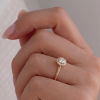 A close-up of a hand wearing a gold engagement ring with a round-cut clear center stone and a halo of small clear stones, set against a soft neutral background.
